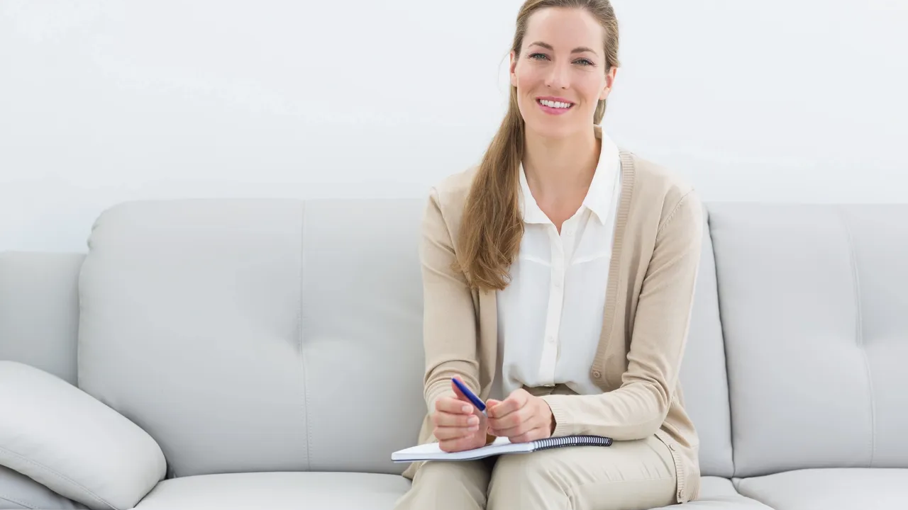 Woman sitting on a white sofa with a notepad in her hand wearing a light brown cardigan and khaki pants