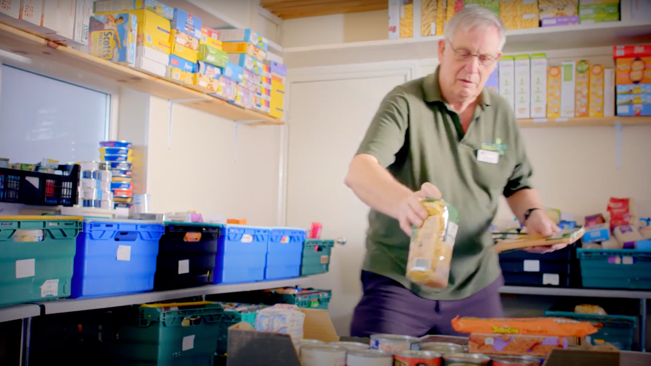 An elderly man sorting through food bank donation bins in a small storage room