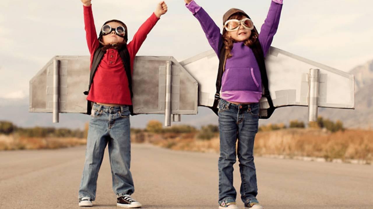 Two children dressed as pilots with goggles and plane wings attached to their backs stand on a wide road on an empty field with their arms raised up high