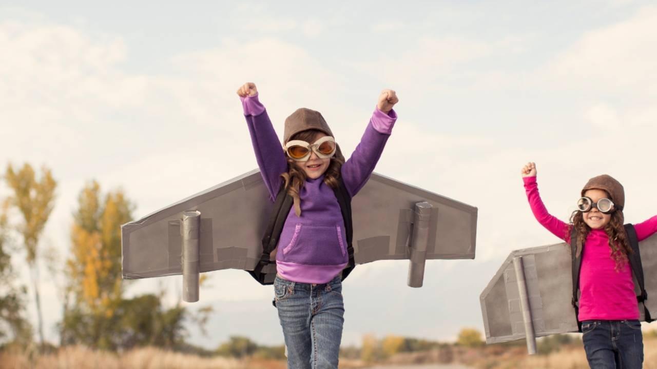 A child wearing a purple sweater with plane wings strapped to her back runs enthusiastically towards the camera