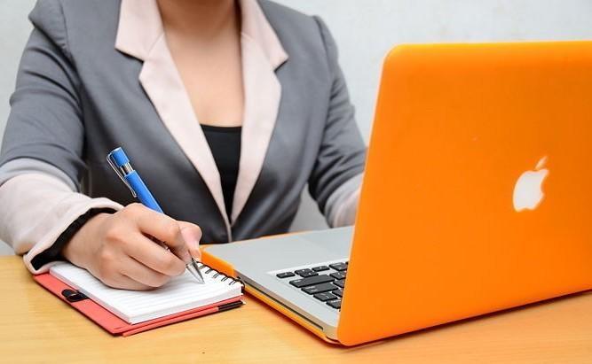 Woman taking notes on her notebook while listening an online workshop on her orange laptop