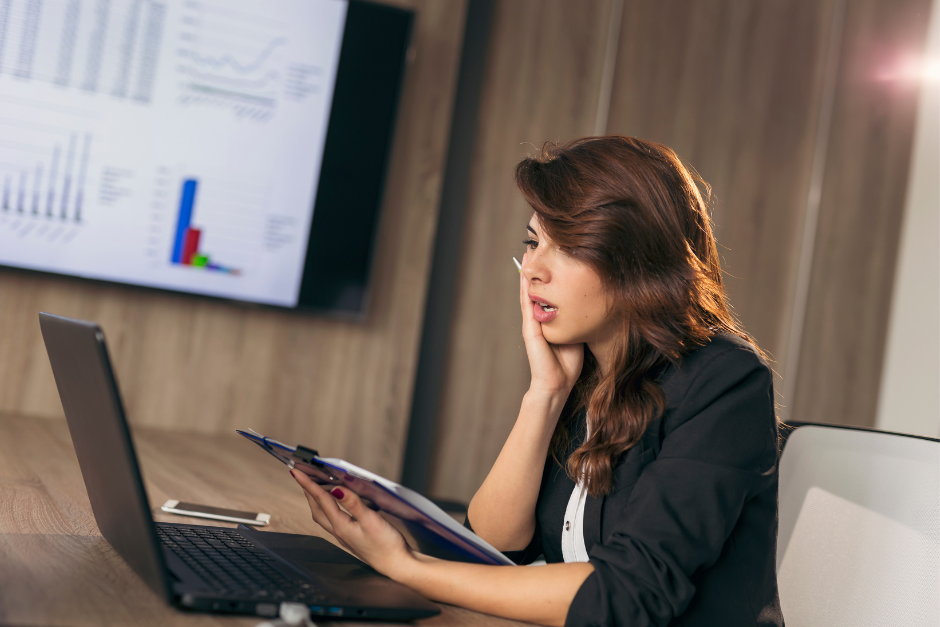 woman at computer looking nervous and anxious holding a clipboard with bookkeeping numbers and charts