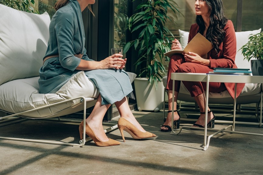 two women are in community together, sharing ideas and supporting one another while they sit on comfortable furniture in a green office